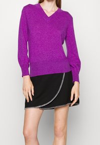 Pull en maille violet avec col en V et manches longues bouffantes, associé à une jupe portefeuille noire avec des coutures blanches le long de l'ourlet.