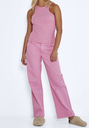 Kvinde iført en ærmeløs lyserød top, løse lyserøde bukser og beige pelsede hjemmesko, stående foran en ensfarvet hvid baggrund.