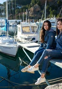 Due persone sono sedute su un molo di legno accanto a delle barche. Indossano giacche con cappuccio blu e jeans, uno in scarpe bianche e l'altro in beige.
