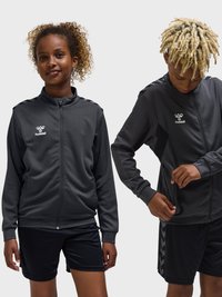 To unge personer iført sorte sportjakker og shorts, den ene vender fremad og smiler, den anden justerer lynlåsen på jakken.