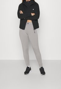Sudadera negra con cremallera y cuello alto, pantalones de chándal ajustados grises, y zapatillas deportivas negras con suelas blancas, exhibidas sobre un fondo liso.