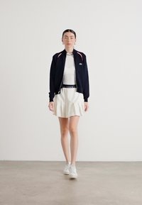 Tamsiai mėlynas zip-up džemperis su raudonomis detalėmis, baltas prigludęs viršus ir klostuota balta sijonas. Modelis vilki baltus sportinius batus. Minimalistinis fonas.