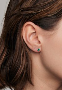 Zielone, szmaragdowe, kwadratowe kolczyki z małymi, przezroczystymi akcentami, osadzone w srebrnym metalu, noszone w płatku ucha obok średnio brązowych włosów.