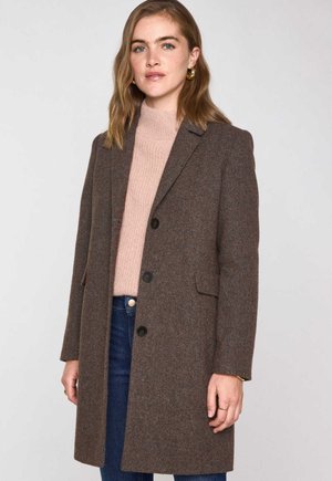 Manteau en chevrons marron avec un col à revers, deux poches avant et trois boutons. Porté sur un pull texturé rose pâle et un jean bleu.