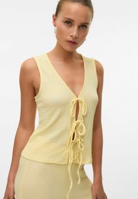 Top giallo senza maniche con scollo a V e rifinitura annodata. Tessuto leggero e strutturato con una vestibilità ampia e un'apparenza leggermente stropicciata.