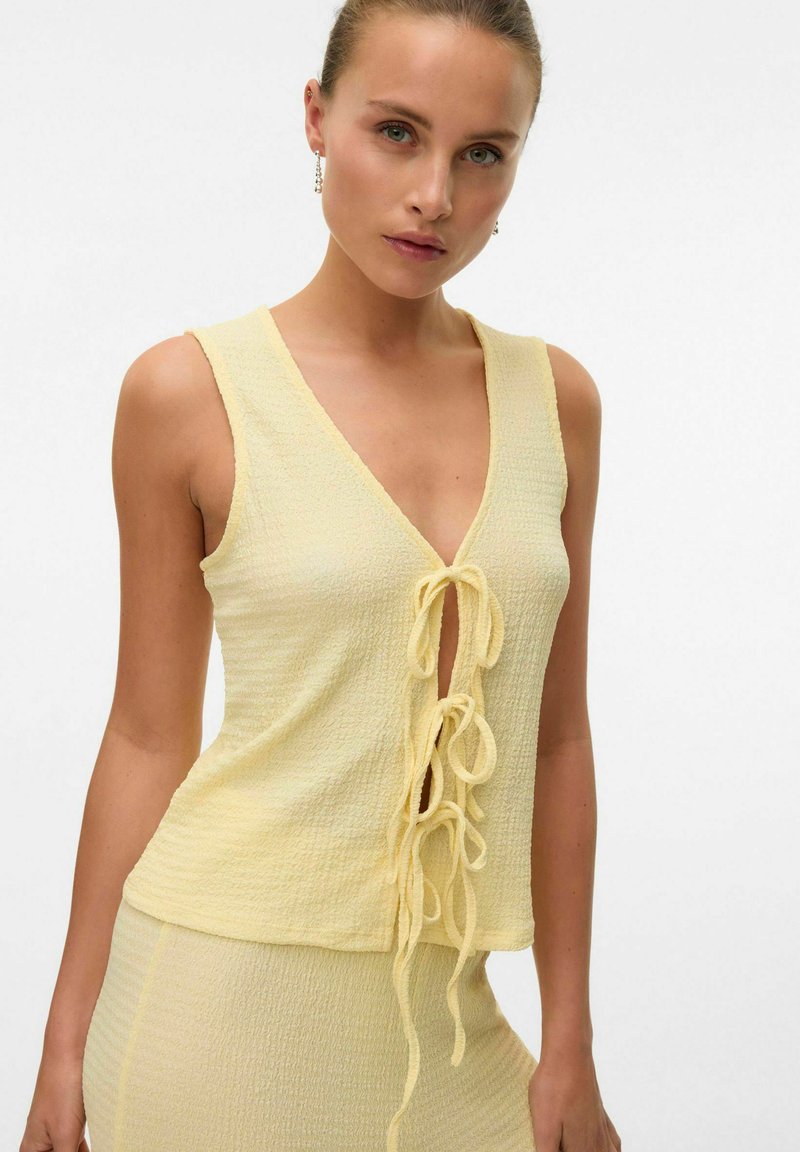 Top giallo senza maniche con scollo a V e rifinitura annodata. Tessuto leggero e strutturato con una vestibilità ampia e un'apparenza leggermente stropicciata.