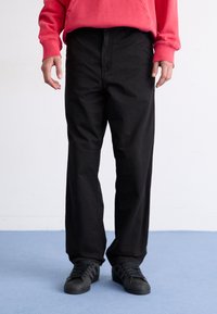Pantalones de algodón negros con un diseño de pierna recta, que cuentan con una cintura alta y bolsillos laterales, combinados con zapatillas negras sobre un suelo azul.