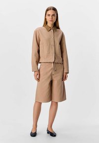 Beige kort jacka med två framfickor, knappstängning och krage; parat med knälånga shorts, båda i liknande tyg och färg.