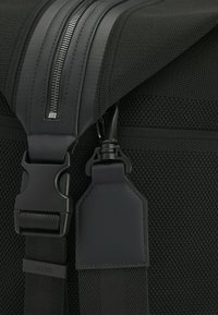 Sac en textile noir avec un extérieur rembourré, fermeture à glissière brillante et bandoulière réglable avec une boucle en plastique robuste et un porte-étiquette.