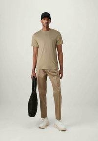 Olivengrønn t-skjorte, beige skreddersydde bukser, hvite joggesko og en svart tote bag, båret av en person som står mot en lys bakgrunn.