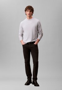 Light grey long-sleeve shirt, fitted at the torso. Black jeans with a slim fit. Black shoes. Neutral gray background. Model standing with hands in pockets.