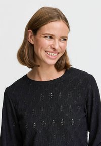 T-shirt noir à manches longues en tissu texturé avec un motif en losanges et de petites découpes, col rond et coupe décontractée.