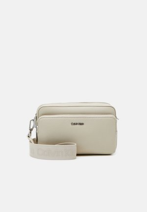 Sac poignet beige Calvin Klein avec deux compartiments zippés et une sangle en tissu amovible avec le nom de la marque gravé.