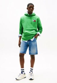 Grüner Kapuzenpullover mit einer Vorderseite-Tasche, kombiniert mit blauen Denim-Shorts. Weiße Sneakers mit marineblauen Akzenten und gestreiften Socken runden den Look ab.
