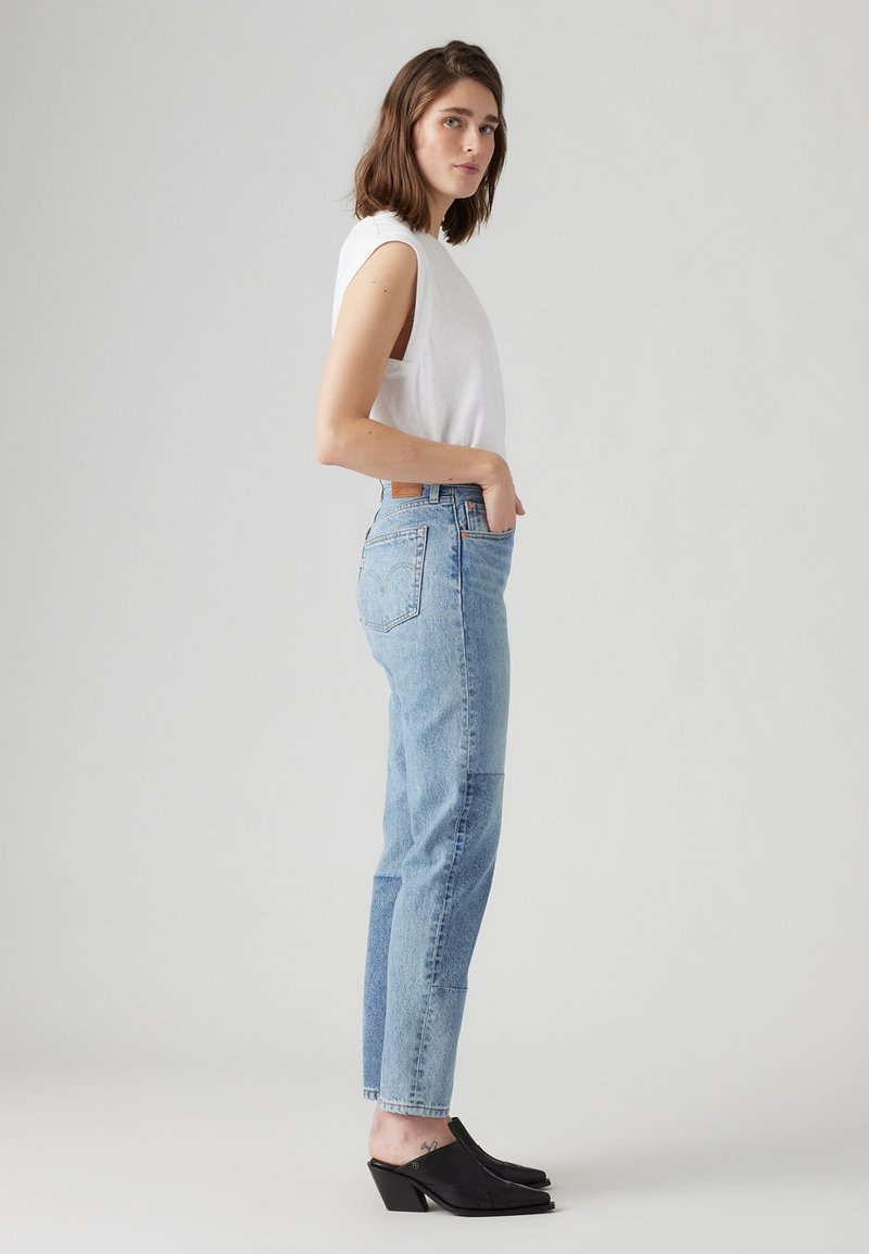 Top bianco senza maniche, jeans azzurri a vita alta con dettagli a toppa, mule nere con tacco a blocco, in piedi su uno sfondo neutro.
