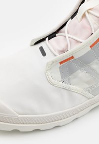 Bílé tenisky s růžovým textilním jazykem, bílými tkaničkami a šedými akcenty zvýrazněnými oranžovým šitím na bílém pozadí.