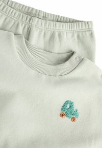 Jasnozielony bawełniany sweatshirt z prążkowanym dekoltem i napami na ramionach, z małym haftowanym turkusowym samochodem z pomarańczowymi kołami. W komplecie z pasującymi spodniami.