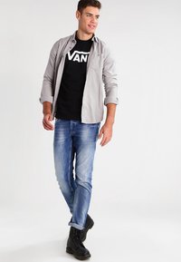 Graues Button-Up-Hemd über einem schwarzen T-Shirt mit "VANS"-Logo, blaue Jeans und schwarze Schnürstiefel. Legere Kleidung mit Details zu hochgerollten Ärmeln.