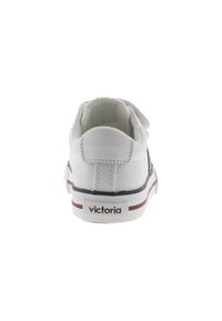 Zapatilla de lona blanca con puntera redonda, correa trasera de Velcro, acentos en azul marino y suela de rayas rojas y blancas; cuenta con el logotipo "victoria" en el talón.