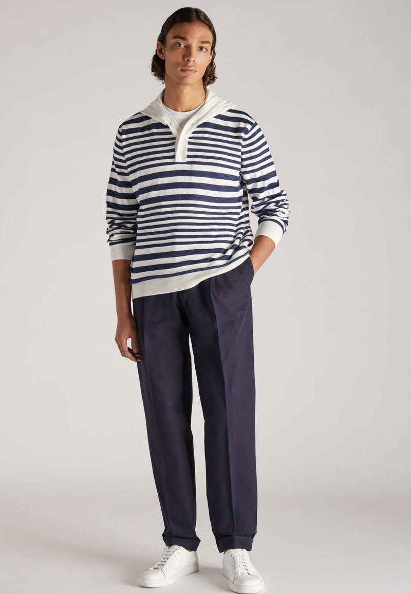 Junger Mann, der mit einer Hand in der Tasche steht, trägt ein gestreiftes langärmliges Oberteil in Weiß und Marineblau, dunkle Hose und weiße Sneaker vor einem einfarbigen Hintergrund.