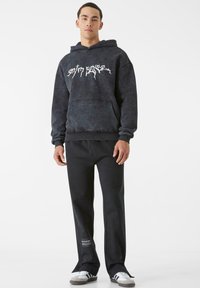 Juoda sportinė striukė su gobtuvu, ant kurios yra balta grafika, derinama su laisvais juodais kelnais su logotipo užklipu; modelis dėvi juodai baltus sportbačius.