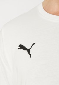 Balta medvilninė marškinėliai su juodu išsiuvinėtu Puma logotipu kairiajame krūtinės gale. Audinys yra lygus ir turi apvalų kaklą.