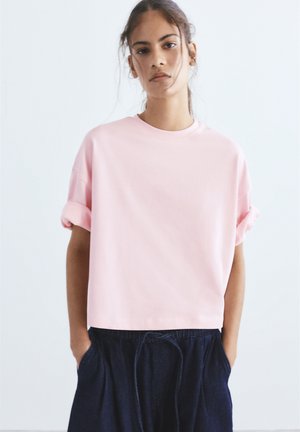 Giovane donna con capelli sciolti che indossa una maglietta rosa chiaro a maniche corte con polsini arrotolati e pantaloni scuri con coulisse, mani nelle tasche, sfondo neutro.