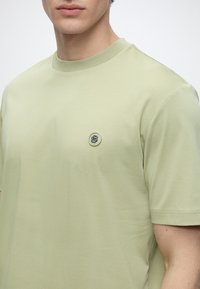 Homme portant un t-shirt vert clair à manches courtes avec un petit logo rond sur la poitrine gauche sur un fond uni.