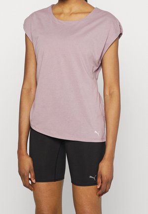 Kobieta ubrana w luźny, jasnofioletowy top bez rękawów i dopasowane czarne szorty Puma, stojąca na jednolitym tle.