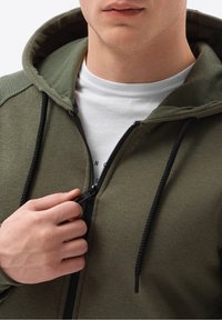 Felpa con cappuccio verde oliva con zip e design testurizzato sulle spalle, zip nera e coulisse. Indossata sopra una maglietta bianca con stampa sotto.