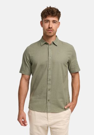 Mann mit kurzen lockigen Haaren, der ein hellgrünes kurzärmeliges Hemd mit Knöpfen und beige Hosen trägt, steht mit einer Hand in der Tasche.