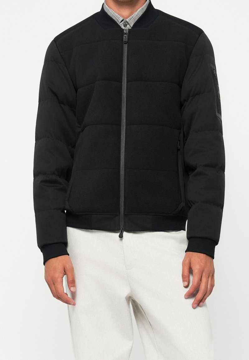 Homme portant une veste bomber noire matelassée avec une fermeture éclair complète et des poignets noirs, associée à un pantalon clair et un col de chemise à carreaux.