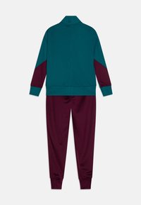 Tuta sportiva con giacca teal e accenti bordeaux, dotata di colletto alto, abbinata a pantaloni bordeaux, entrambi realizzati in un materiale liscio e elastico.
