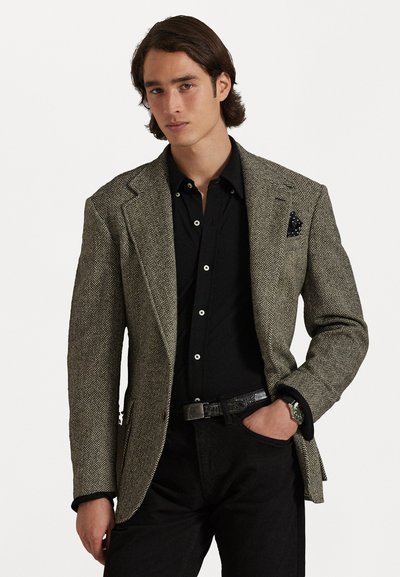 Blazer u houndstooth uzorku u smeđoj i crnoj boji, s naglašenim ramenima, džepom na prsima sa tamnim uzorčanim maramom, uparen s crnom košuljom.