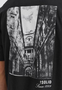 T-shirt en coton noir avec un imprimé architectural monochrome représentant une vue d'une rue étroite, des détails de bâtiments et le texte "!SOLID Since 1999."