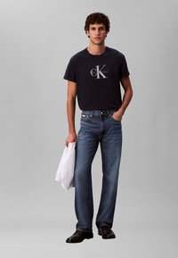 Tmavomodré bavlnené tričko s bielym logom Calvin Klein, doplnené svetlomodrými rovnými džínsami. Model drží bielu bundu a má obuté čierne topánky.