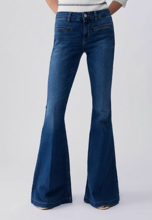 Mujer con vaqueros de campana azul oscuro de cintura alta, con cremallera frontal y bolsillos horizontales con aberturas, sobre un fondo liso.