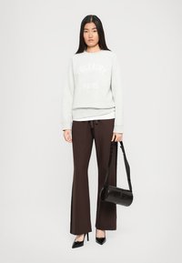 Femme portant un pull gris clair de Voltaire Paris, un pantalon marron foncé, des escarpins noirs à talons aiguilles, tenant un sac à main cylindrique noir.