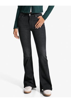 Femme portant un jean noir évasé taille haute avec poches avant, associé à des baskets blanches et un top à manches longues vert foncé.