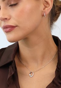 Silberne Halskette mit einem ovalen Anhänger, der einen funkelnden Rand aus Kristallen hat; getragen mit einem braunen Satinhemd und Ohrreifen.