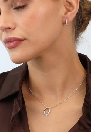 Silberne Halskette mit einem ovalen Anhänger, der einen funkelnden Rand aus Kristallen hat; getragen mit einem braunen Satinhemd und Ohrreifen.