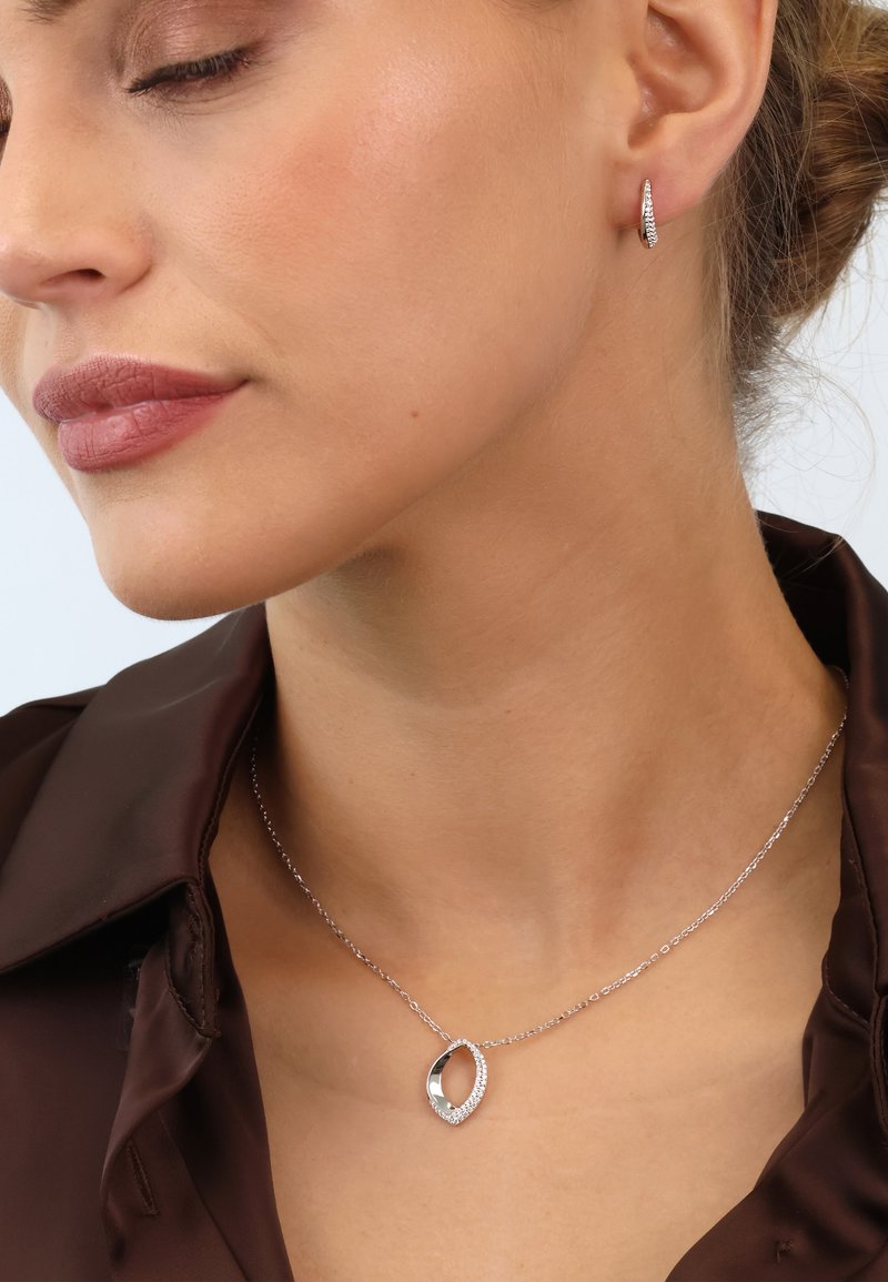 Silberne Halskette mit einem ovalen Anhänger, der einen funkelnden Rand aus Kristallen hat; getragen mit einem braunen Satinhemd und Ohrreifen.