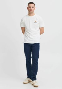 Hvit bomulls t-skjorte med en liten brun logo på venstre bryst, kombinert med mørkeblå denimjeans og hvite joggesko med beige detaljer.