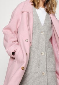 Rosa Wollmantel mit Kragen und abgerundeten Knöpfen, getragen über einer grauen Rippstrick-Strickjacke mit V-Ausschnitt und zwei Vorderentaschen.