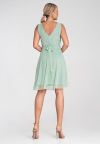 Miętowa sukienka z dekoltem w kształcie V, bez rękawów, z paskiem w talii i zwiewnym, przezroczystym dołem. W połączeniu z srebrnymi, przewiązanymi szpilkami.
