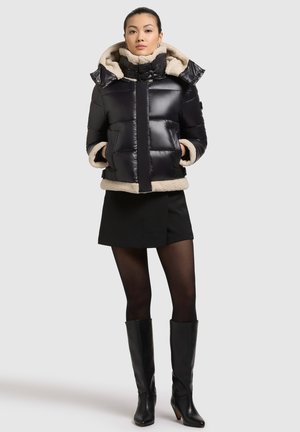 Giacca a vento nera lucida con bordo in fleece beige, chiusura con zip, grande cappuccio e due tasche frontali. Indossata sopra una gonna nera e collant.