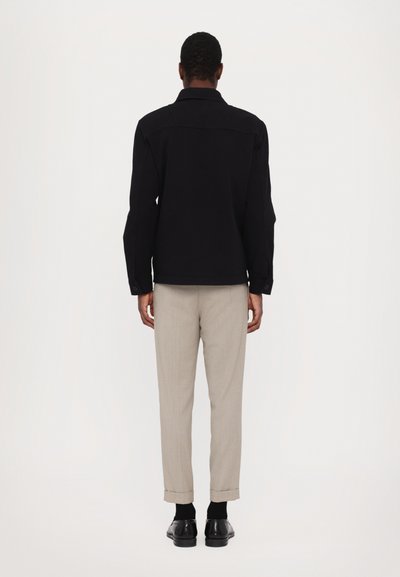 Black button-up jacket with a straight cut, paired with light beige trousers and black shoes. Smooth fabric with a tailored fit.
