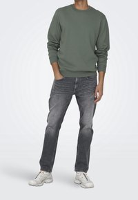 Sudadera de cuello redondo verde, jeans grises desgastados y zapatillas blancas. Ajuste casual con puños y bajo de canalé. Textura suave, detalles sutiles de desgaste.