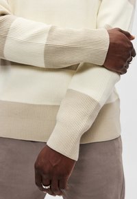 Beiger und cremefarbener gestreifter Strickpullover mit gerippter Textur. Verfügt über kontrastierende Farbabsetzungen an Ärmeln und Bündchen.