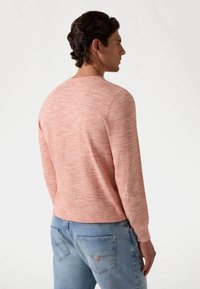 Pulover lung din tricot de culoare corai deschis, cu mâneci lungi și guler rotund, având un model texturat, purtat cu blugi albaștri deschisi.